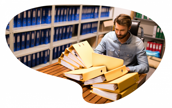 Numen propose une gamme complète de services archivistiques sur mesure. Organisez, sécurisez et valorisez vos documents avec Nuemn Prestations Archivistiques. Image d'un homme assis dans une salle d'archives. On y voit des classeurs remplis de documents sur des étagères. Une pile de classeurs est entassée devant lui.