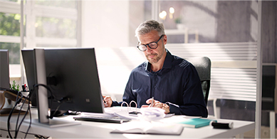 Numen propose un accompagnement stratégique pour vos archives dans le but d'identifier vos besoins stratégiques et défiinr une gouvernance documentaire rapide. Image d'un homme assis à son bureau devant un ordinateur. Il regarde des documents papier placés devant lui